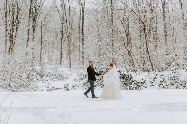 Beautiful wedding couple walking in winter snowy forest, woman in white dress and poncho, bearded man in black coat