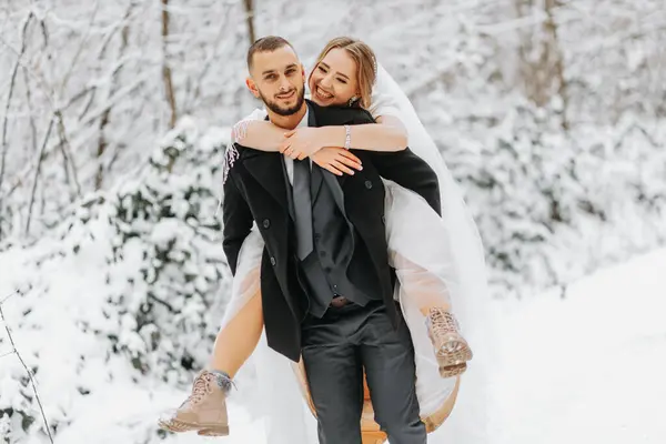 Beautiful wedding couple walking in winter snowy forest, woman in white dress and mink fur coat, bearded man in black coat. The groom carries the bride in his arms, a funny photo