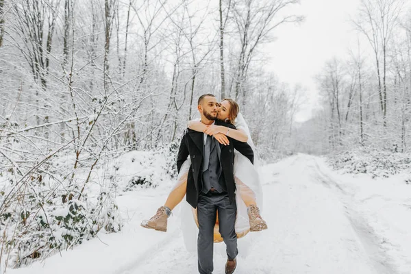 Beautiful wedding couple walking in winter snowy forest, woman in white dress and mink fur coat, bearded man in black coat. The groom carries the bride in his arms, a funny photo