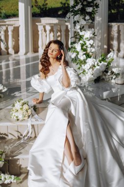 Çiçeklerin arka planında güzel bir gelin, dışarıda oturan şık gelinlikli çekici bir kadın. Mutlu gelin kadın. Kıvırcık uzun saçlı gelin. Düğün günü. Evlilik..