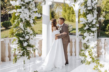 Zarif bir gelin ve damat, güneşli bir düğün gününde güzel bir yerde çiçeklerin arka planına karşı birlikte poz verirler. Damat geline nazikçe sarılır ve onu öper..