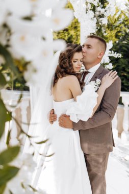 Zarif bir gelin ve damat, güneşli bir düğün gününde güzel bir yerde çiçeklerin arka planına karşı birlikte poz verirler. Damat geline nazikçe sarılır ve onu öper..