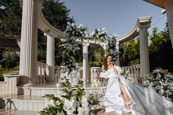 花の背景に美しい花嫁 屋外に座っているファッショナブルなウェディングドレスの魅力的な女性 ハッピーな花嫁さん カーリーロングヘアの花嫁 結婚式の日 結婚について — ストック写真
