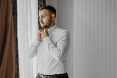 Yakışıklı bir adam sabahleyin bir otel odasında pencerede gömlek giyiyor. Bir etkinlik ya da yeni bir iş günü için hazırlık. Yeni fırsatlar, tanıdıklar, düğün günü veya röportaj hazırlığı