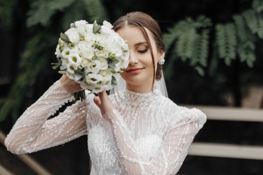 Taşlar ve boncuklarla işlenmiş beyaz gelinlik içinde gelin bir buket gül ve yeşillik taşıyor. Şık bir düğün buketi.