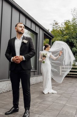 Şık bir elbise ve duvak giymiş gelin. Ön planda siyah takım elbiseli damat. Güzel bir düğün konsepti.