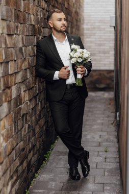 Şık ve zarif bir genç adam. Siyah takım elbiseli ve beyaz gömlekli. Elinde çiçek demeti var.