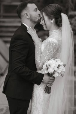 Damat geline nazikçe sarılır ve dudaklarıyla alnına dokunur, gelin ellerini damadın omuzlarına koyar ve anın tadını çıkarır. Siyah beyaz fotoğraf
