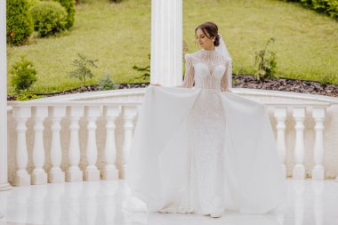 Beyaz bir gelinlik içinde kolları olan genç bir gelinin tam boy portresi. Profesyonel saç stili ve doğal makyaj. Güzel bir yerde düğün kavramı.