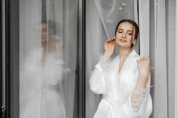 portrait of bride girl in white gown with professional hairstyle and natural makeup in hotel room with reflection in window. Wedding dress with sleeves on a mannequin. The best day