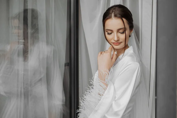 portrait of bride girl in white gown with professional hairstyle and natural makeup in hotel room with reflection in window. Wedding dress with sleeves on a mannequin. The best day