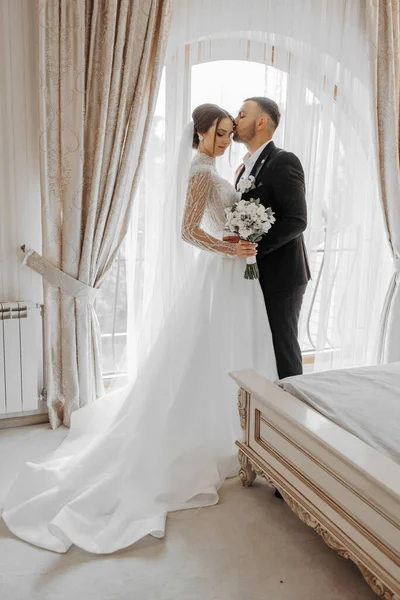 a joyful couple of young brides hug, kiss in a hotel room. The bride in a wedding dress hugs the cheerful groom in classic formal clothes by the shoulder. The first meeting of the bride and groom