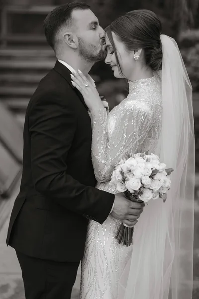 The groom gently hugs the bride and touches her forehead with his lips, the bride puts her hands on the groom\'s shoulders and enjoys the moment. Black and white photo