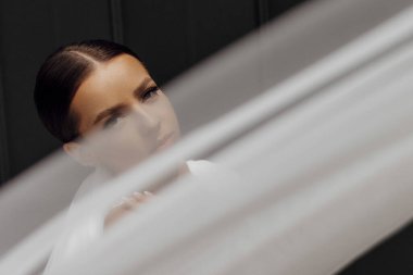 Güzel gelin kız uzun peçeli beyaz elbise giyiyor ve modern şehrin açık havada, güneş ışığı altında gri duvarın yanında poz veriyor..