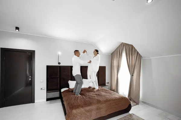 The morning of the bride and groom in a hotel room. Happy and in love bride and groom jumping and having fun on the bed. Confident girl-bride and handsome groom. Preparation for the wedding