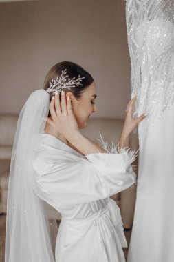 İpek takım elbiseli ve peçeli bir gelin odasında gelinliğinin yanında poz veriyor. Gelinin sabahı. Hazırlık. Düğün fotoğrafçılığı