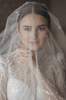 Düğün portresi. Gelin zarif bir gelinlik içinde duvağa sarılmış pozlar veriyor. Gelinin sabahı. Güzel saç ve makyaj.