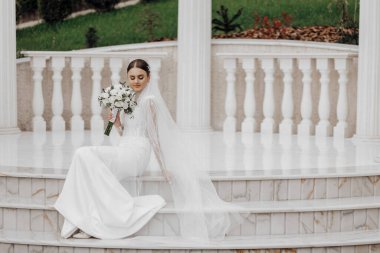 Uzun elbiseli gelin, beyaz Roma tarzı sütunların yanında oturuyor. Güzel saç ve makyaj. Nefis bir düğün.