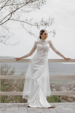 Portre. Şık, uzun bir elbise ve peçeyle gelin elinde buket, ahşap korkulukların yanında poz veriyor. Doğada düğün