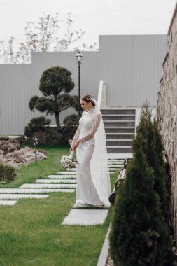 Portre. Gelin zarif, uzun elbiseli ve peçeli, elinde buket, bahçede poz veriyor. Doğada düğün