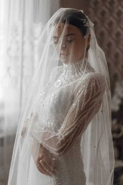Wedding portrait. The bride in an elegant wedding dress poses wrapped in a veil. Morning of the bride. Beautiful hair and makeup