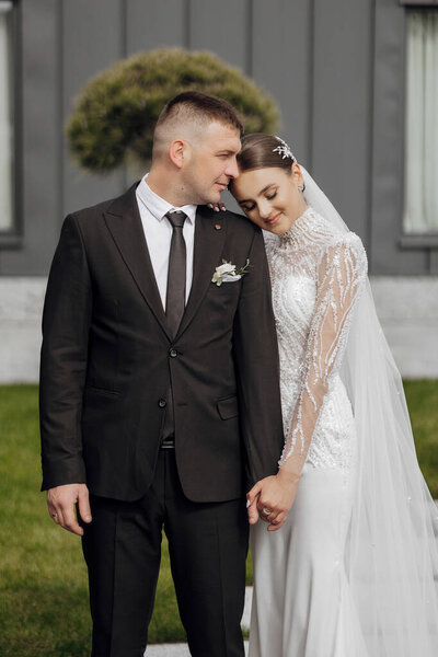 Портрет. Невеста в элегантном длинном платье и жених обнимаются и позируют во дворе, стоя на каменной дорожке. Свадьба на природе