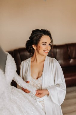 Dantel bir elbise ve peçeli ipek bir bornoz giyen bir gelin odasında gelinliğinin yanında poz veriyor. Gelinin sabahı. Hazırlık. Düğün fotoğrafçılığı