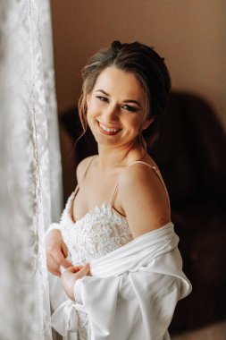 Dantel bir elbise ve peçeli ipek bir bornoz giyen bir gelin odasında gelinliğinin yanında poz veriyor. Gelinin sabahı. Hazırlık. Düğün fotoğrafçılığı