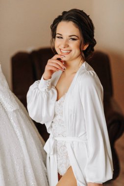 Dantel bir elbise ve peçeli ipek bir bornoz giyen bir gelin odasında gelinliğinin yanında poz veriyor. Gelinin sabahı. Hazırlık. Düğün fotoğrafçılığı