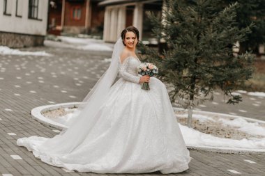 Trenli ve uzun peçeli beyaz elbiseli gelin, elinde buketle doğada poz veriyor. Yürü hadi. Doğada kış düğünü.