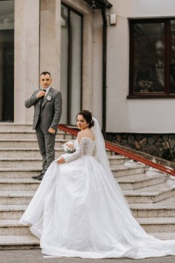 Beyaz elbiseli, trenli bir gelin ve takım elbiseli bir damat binanın merdivenlerinde poz veriyor. Doğada düğün fotoğrafı çekimi