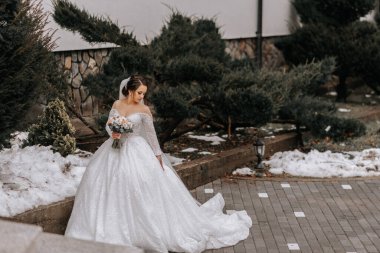 Trenli ve uzun peçeli beyaz elbiseli gelin, elinde buketle doğada poz veriyor. Yürü hadi. Doğada kış düğünü.