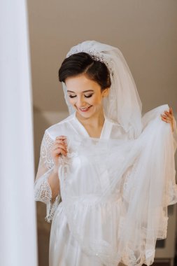 İpek takım elbiseli gelin odasında poz veriyor, peçeyle kendini örtüyor. Gelinin sabahı. Hazırlık. Düğün fotoğrafçılığı