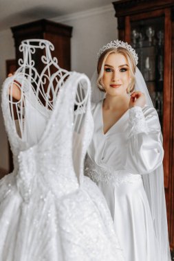 İpek takım elbiseli ve peçeli bir gelin odasında gelinliğinin yanında poz veriyor. Gelinin sabahı. Hazırlık. Düğün fotoğrafçılığı