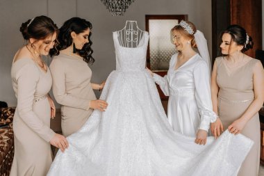 Beyaz cüppeli gelin ve krem rengi elbiseli arkadaşları gelinin elbisesinin yanında poz veriyorlar. Gelinin sabahı