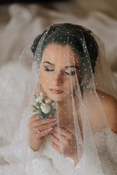 Wedding portrait. The bride in an elegant wedding dress poses wrapped in a veil. Morning of the bride. Beautiful hair and makeup