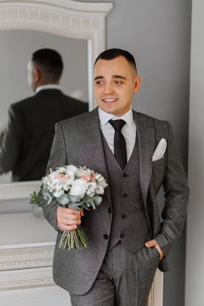 A man in a gray suit is holding a bouquet, posing in his room. Portrait of the groom. Fashion and style. Business