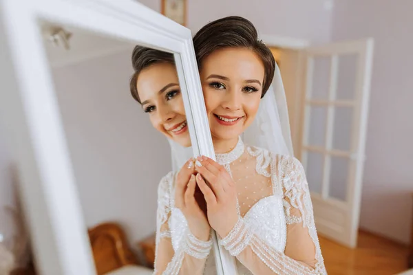 A bride in an elegant dress with long sleeves poses by a mirror in her room.