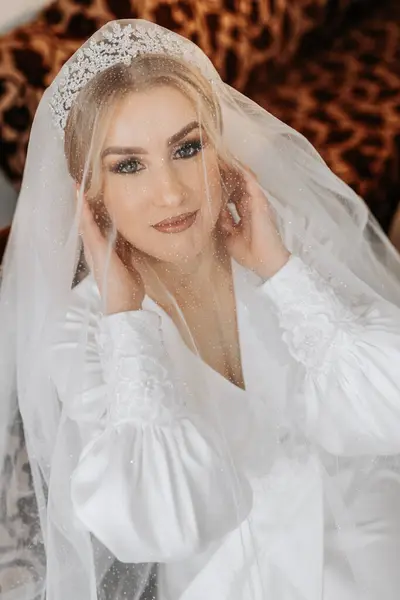 Wedding portrait. Blonde bride in a silk suit and tiara poses behind a sheer veil while sitting in a chair in her room
