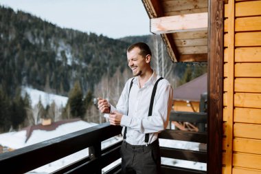 Pantolon askısı takmış, balkonda kar kaplı dağları gören bir saksağanın düğmelerini ilikleyen genç bir adam. Dağlarda dinlenin. Dağ havası. Kışın düğün