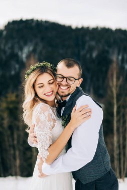 Kış ormanının arka planında mutlu ve gülümseyen gelin ve damat. Yakışıklı bir damat nazikçe gelinine sarılır. Modern kış düğünü konsepti..