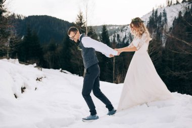 Güzel bir çift kış çam ormanında yürüyor. Etrafı karla çevrili. Beyaz gelinlik giymiş gelin, beyaz gömlek ve yelek giymiş damat. Kış düğünü konsepti.