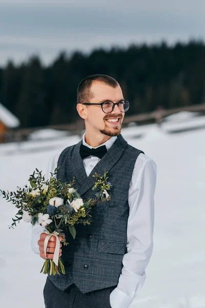 Wedding portrait of the groom. The groom stands against the background of the winter forest. A man in a vest and white shirt, glasses and a bow tie. Winter wedding