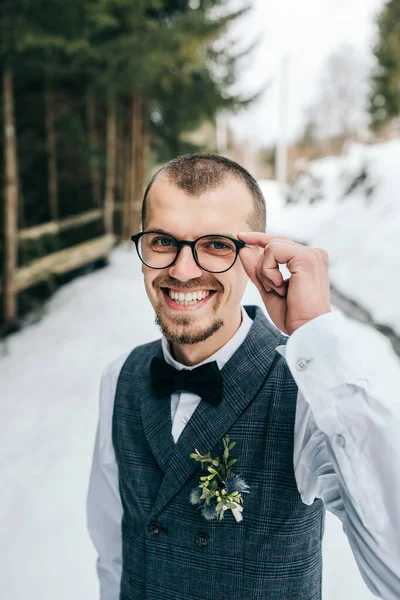 Wedding portrait of the groom. The groom stands against the background of the winter forest. A man in a vest and white shirt, glasses and a bow tie. Winter wedding