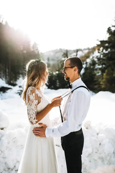 Portrait of the bride and groom against the background of a pine forest and backlight. The bride in a white wedding dress, the groom in a white shirt and trousers with suspenders. Winter wedding.