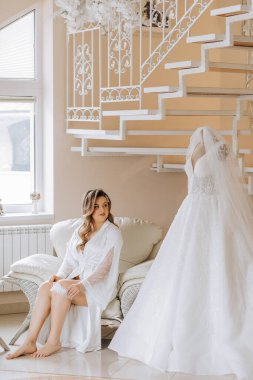 Gelinin düğünden önceki sabahı. Duvağı ve beyaz bornozlu güzel bir saç stili olan güzel bir genç kadın. Gelinliğinin yanında bir mankenin üzerinde. Profesyonel makyaj. Son hazırlıklar.