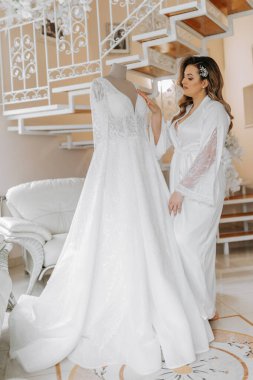 Gelinin düğünden önceki sabahı. Duvağı ve beyaz bornozlu güzel bir saç stili olan güzel bir genç kadın. Gelinliğinin yanında bir mankenin üzerinde. Profesyonel makyaj. Son hazırlıklar.