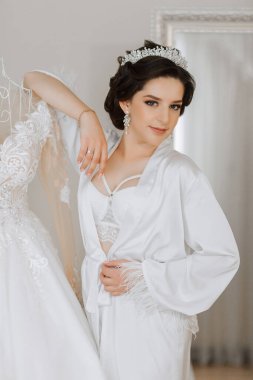 Gelinin düğünden önceki sabahı. Duvağı ve beyaz bornozlu güzel bir saç stili olan güzel bir genç kadın. Gelinliğinin yanında bir mankenin üzerinde. Profesyonel makyaj. Son hazırlıklar.