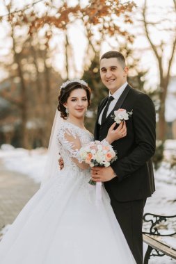 Kış düğünü. Düğün kıyafetleriyle yürüyen mutlu çift, düğün günlerinde kar altında kış parkında sarılıp gülümsüyor. Karlı kış havasında güzel bir çiftin kış aşkı hikayesi.