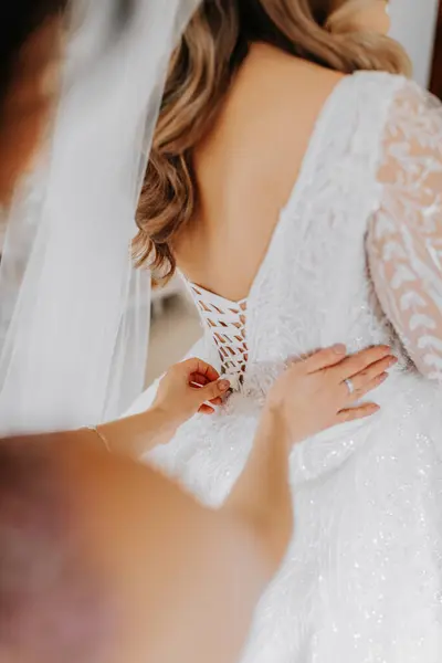 A friend helps the bride fasten her dress. A woman helps her friend fasten the buttons on the back of her wedding dress. The morning of the bride, the creation of a family, an important event.